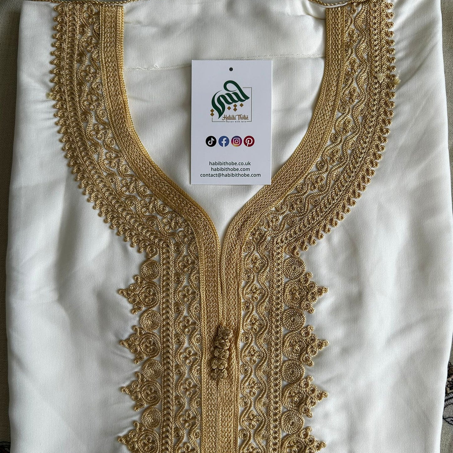 “White Moroccan Gandoura with intricate gold embroidery and tassel detail, folded neatly with branded tag.”