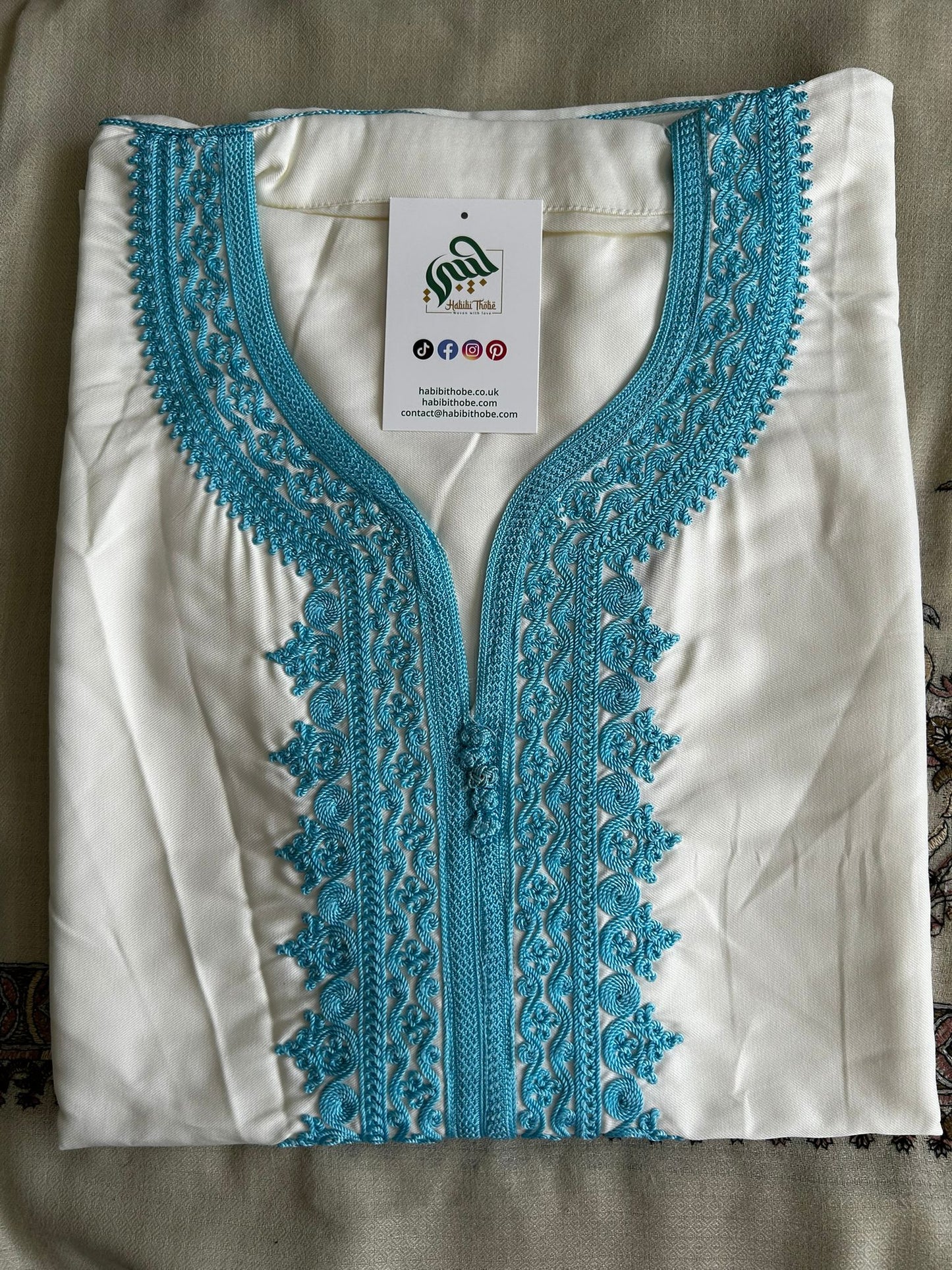 “White Moroccan Gandoura with detailedlight blue embroidery and centralbuttons showcasing refined craftsmanship.”
