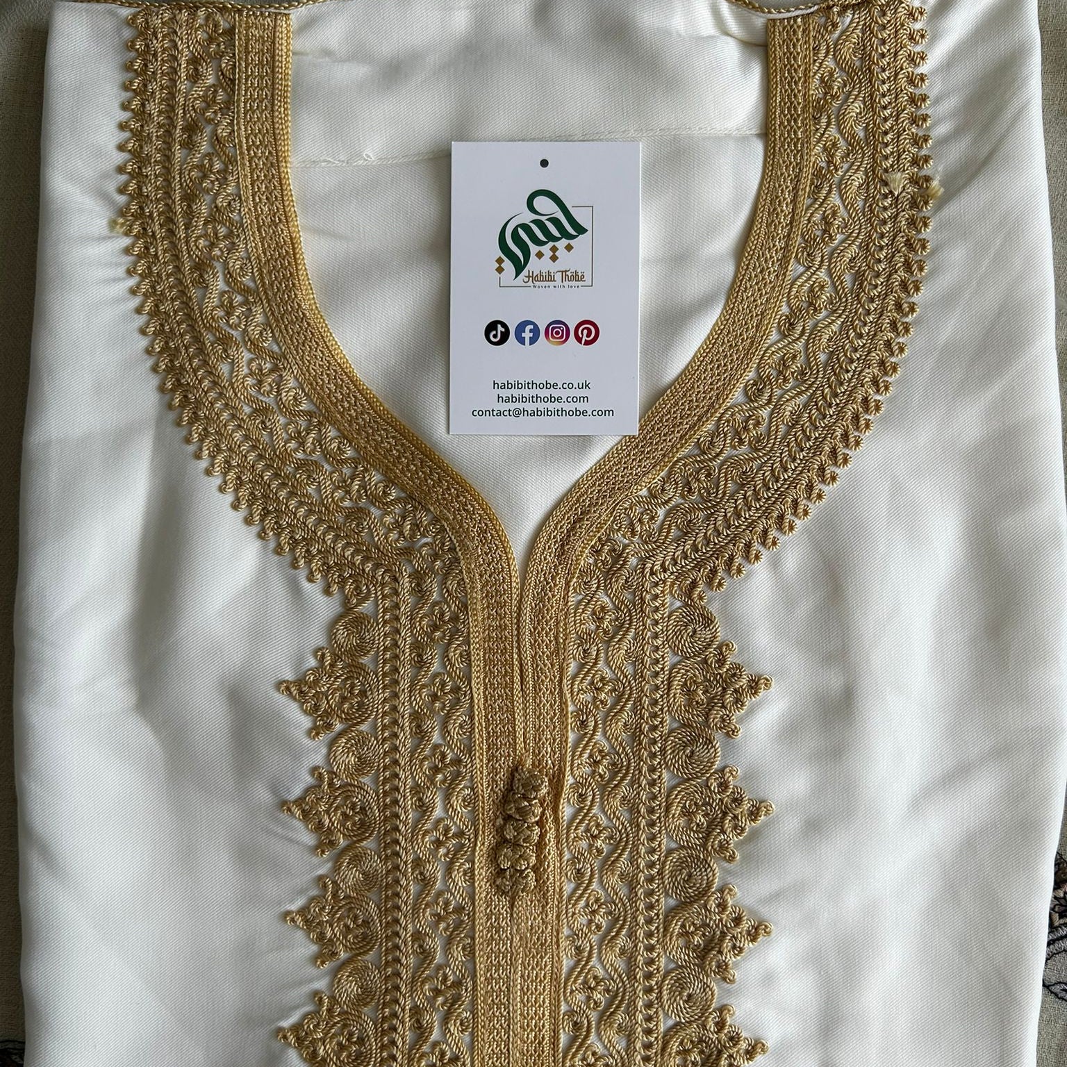 “White Moroccan Gandoura with intricate gold embroidery and tassel detail, folded neatly with branded tag.”