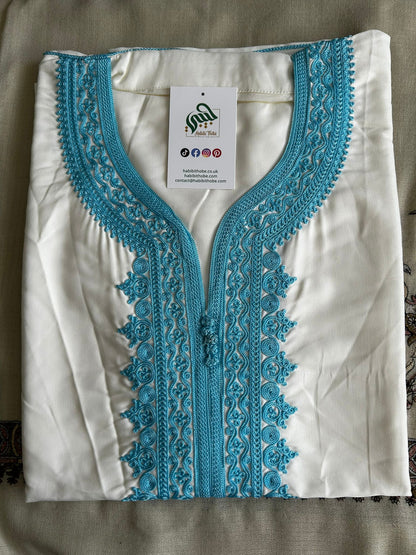  “White Moroccan Gandoura with detailedlight blue embroidery and centralbuttons showcasing refined craftsmanship.”